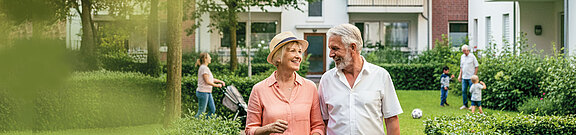 Älteres Paar geht lächelnd durch eine Wohnanlage; im Hintergrund sind moderne Mehrfamilienhäuser und spielende Kinder zu sehen.