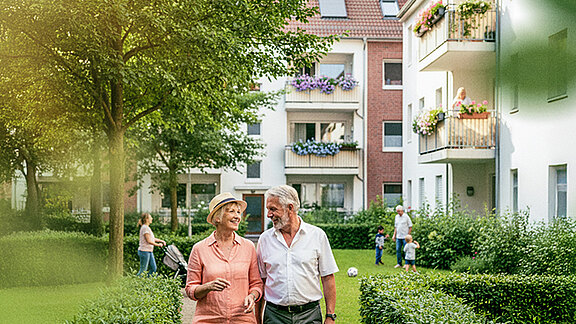 Älteres Paar spaziert lächelnd durch eine begrünte Wohnanlage, im Hintergrund moderne Mehrfamilienhäuser und spielende Kinder.