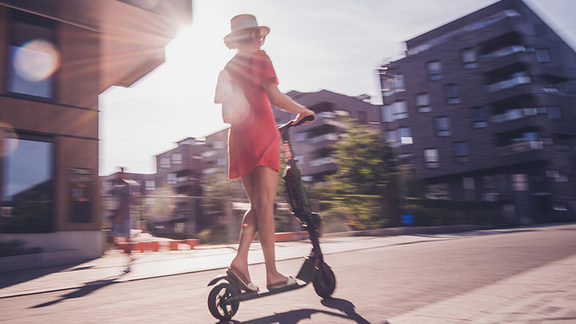 Sommerliche Szene mit Frau auf E-Scooter in einem urbanen Wohnviertel mit Mehrfamilienhäusern.