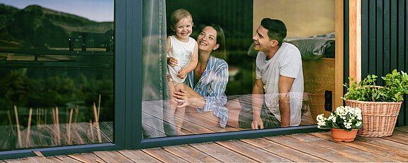 Familie mit kleinem Kind schaut lächelnd aus einem großen Fenster eines modernen Hauses.