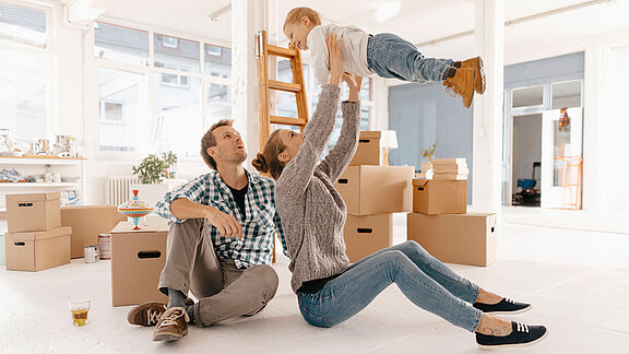 Junge Familie sitzt zwischen Umzugskartons in einer hellen Wohnung; die Mutter hebt lachend ihr Kleinkind in die Luft, der Vater sitzt daneben.