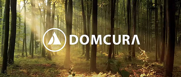 DOMCURA-Logo über einem sonnendurchfluteten Wald mit hohen, schlanken Bäumen.